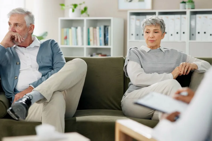 An elderly man and woman sitting on a couch looking distant and contemplative during a therapy session with a professional taking notes.
