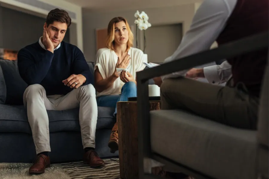 A couple sitting on a couch looking distressed and talking to a therapist in an office setting.