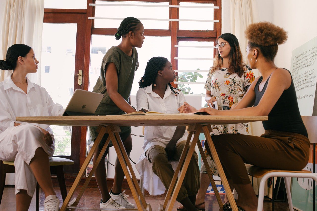 Group of five diverse women having a discussion around a wooden table in a bright room.