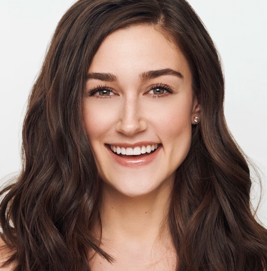 Smiling young woman with long wavy brown hair and clear skin looking at camera on white background.