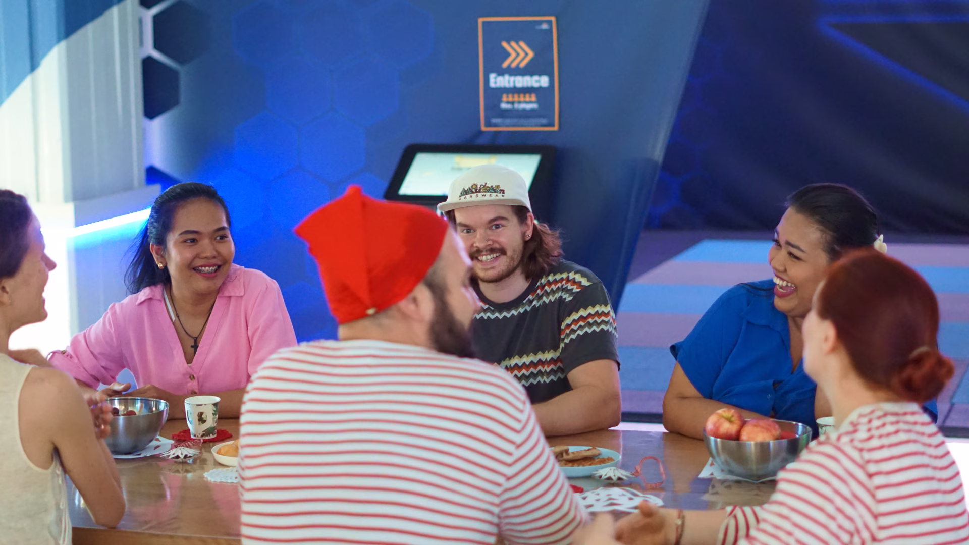A group of adults in christmas outfits sitting around a table and laughing. ValoArena in the background.