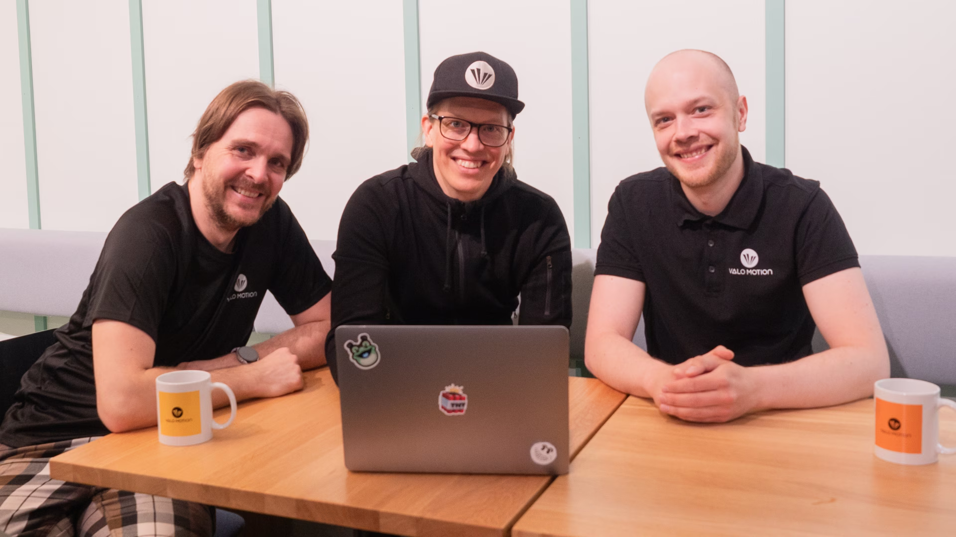 Valo Motion's three founders sitting around a table with an open laptop, smiling.