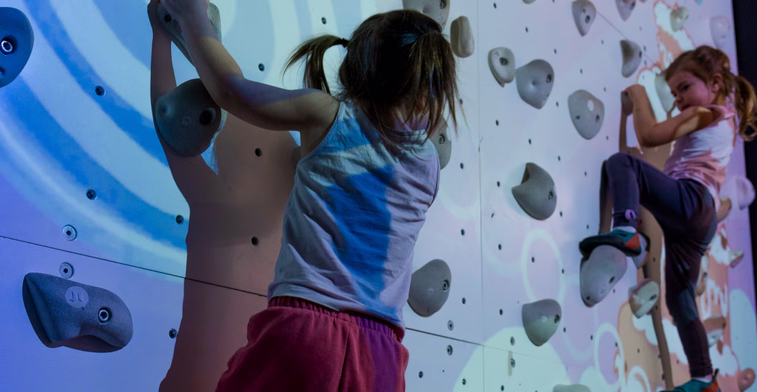 Two small kids playing whack-a-bat on ValoClimb, wearing climbing shoes
