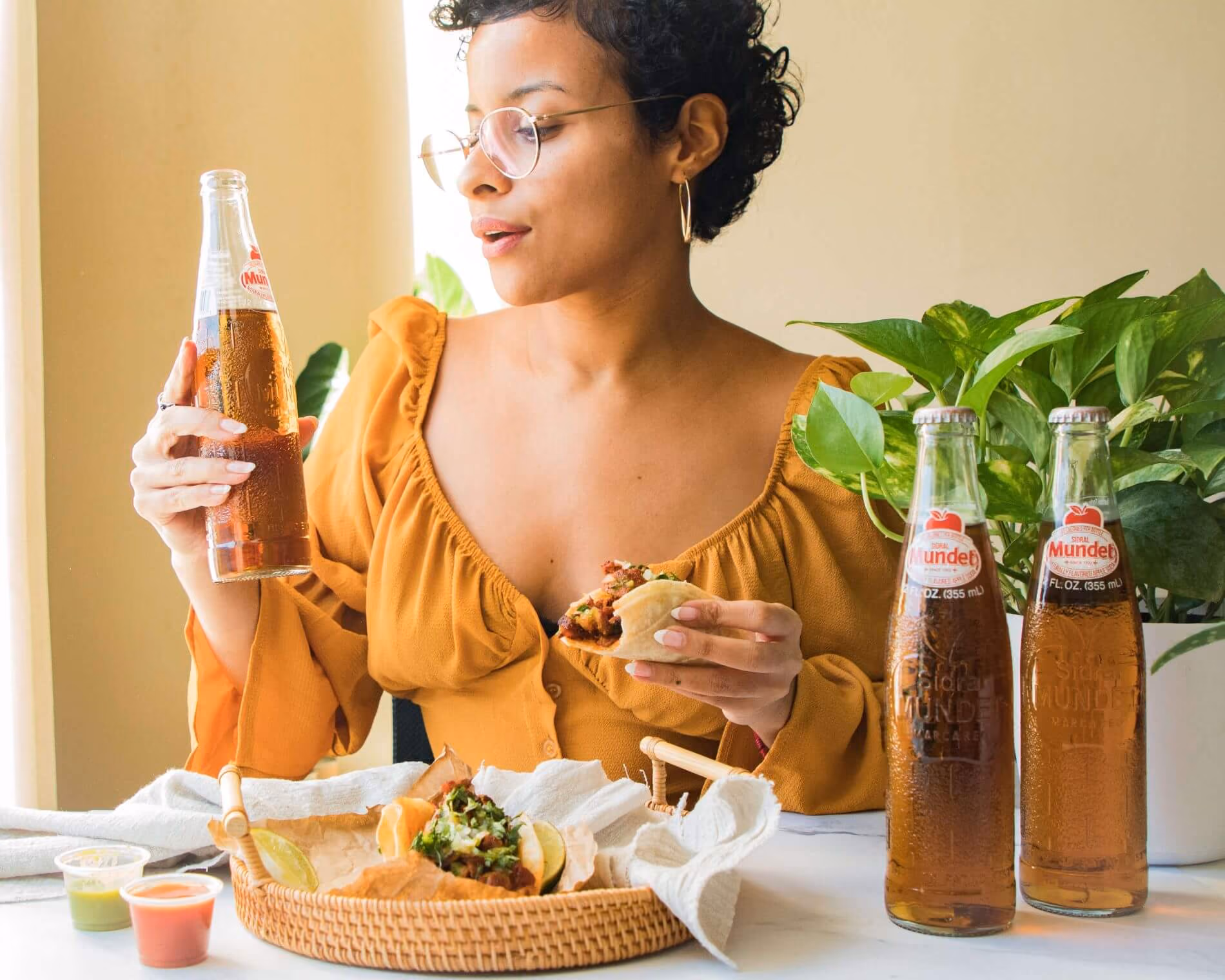 Woman enjoying tacos and a bottle of Sidral Mundet apple soda at a table, with two additional bottles and green plants beside her.