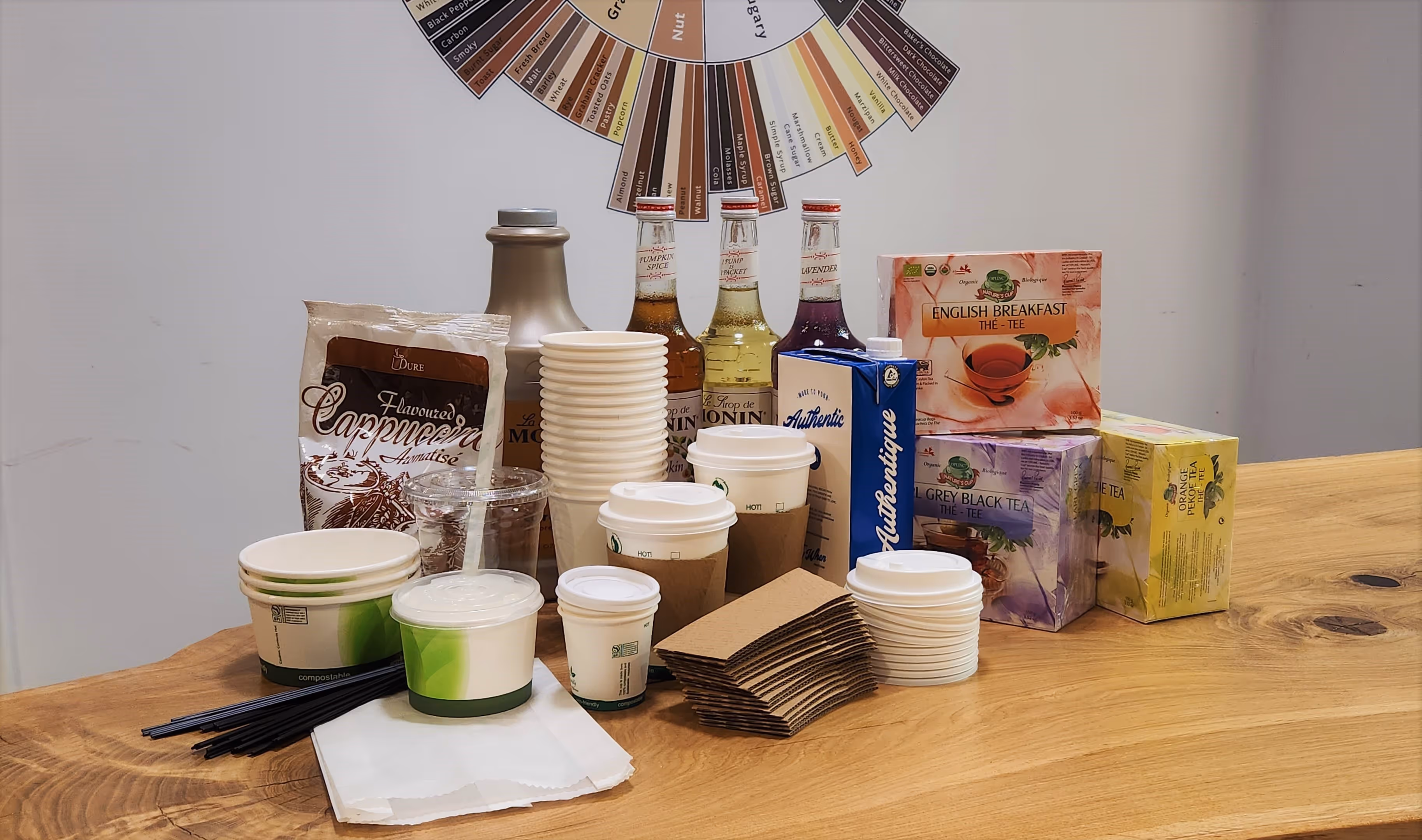 Arrangement of coffee equipment like syrups, paper cups, stirrers, and tea on a wooden table