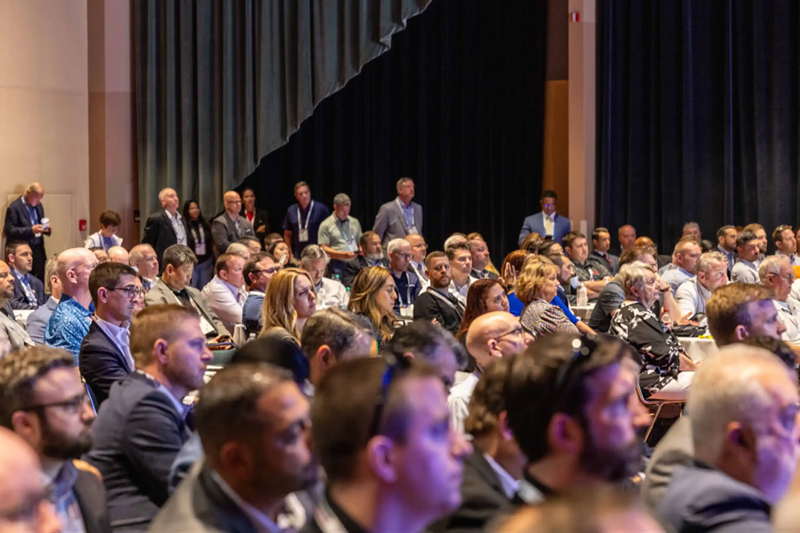 Audience members attentively listening at a formal event, seated at round tables with name tags.