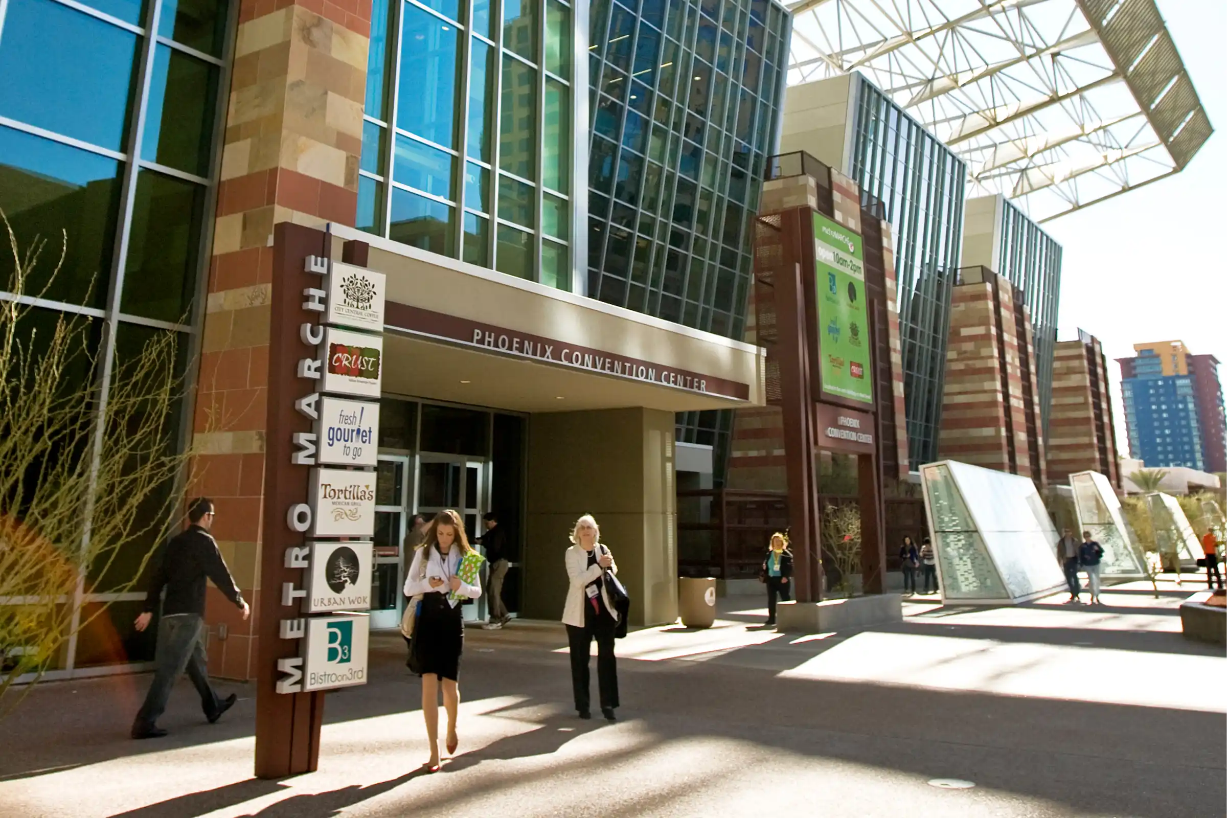 People walking outside the glass-fronted Phoenix Convention Center with Metro Marche restaurant signs visible.