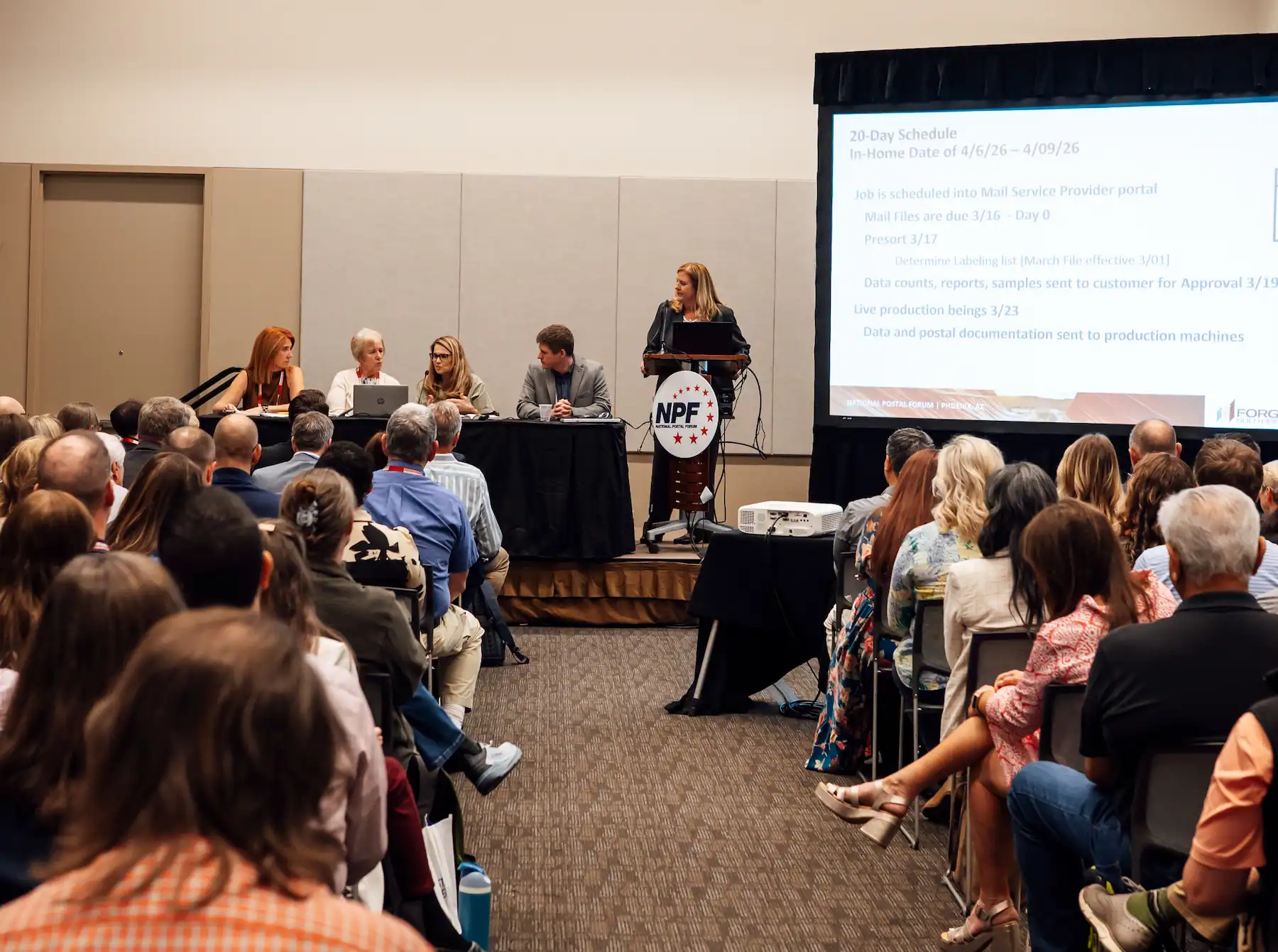 Audience members attentively listening at a formal event, seated at round tables with name tags.
