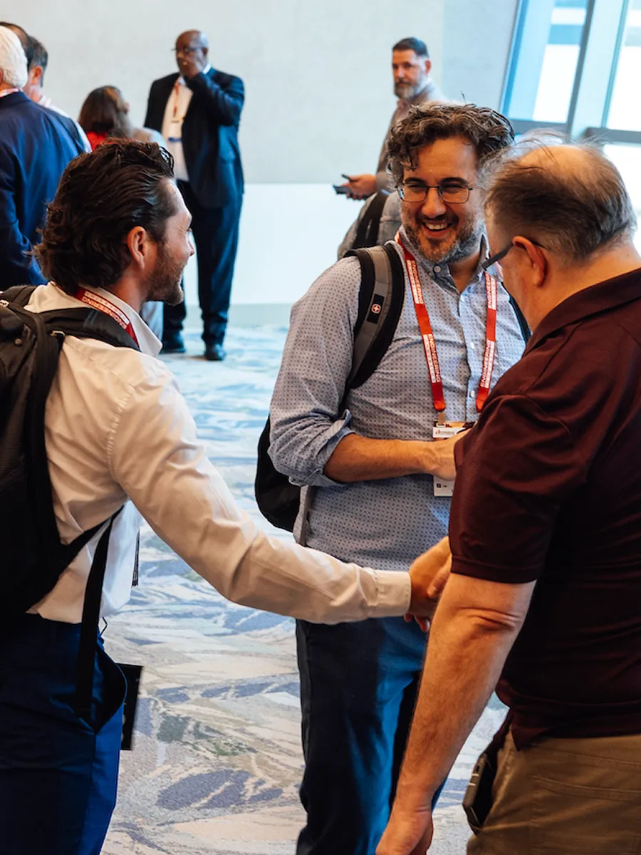Three men in lanyards shaking hands and smiling at a conference or networking event indoors.