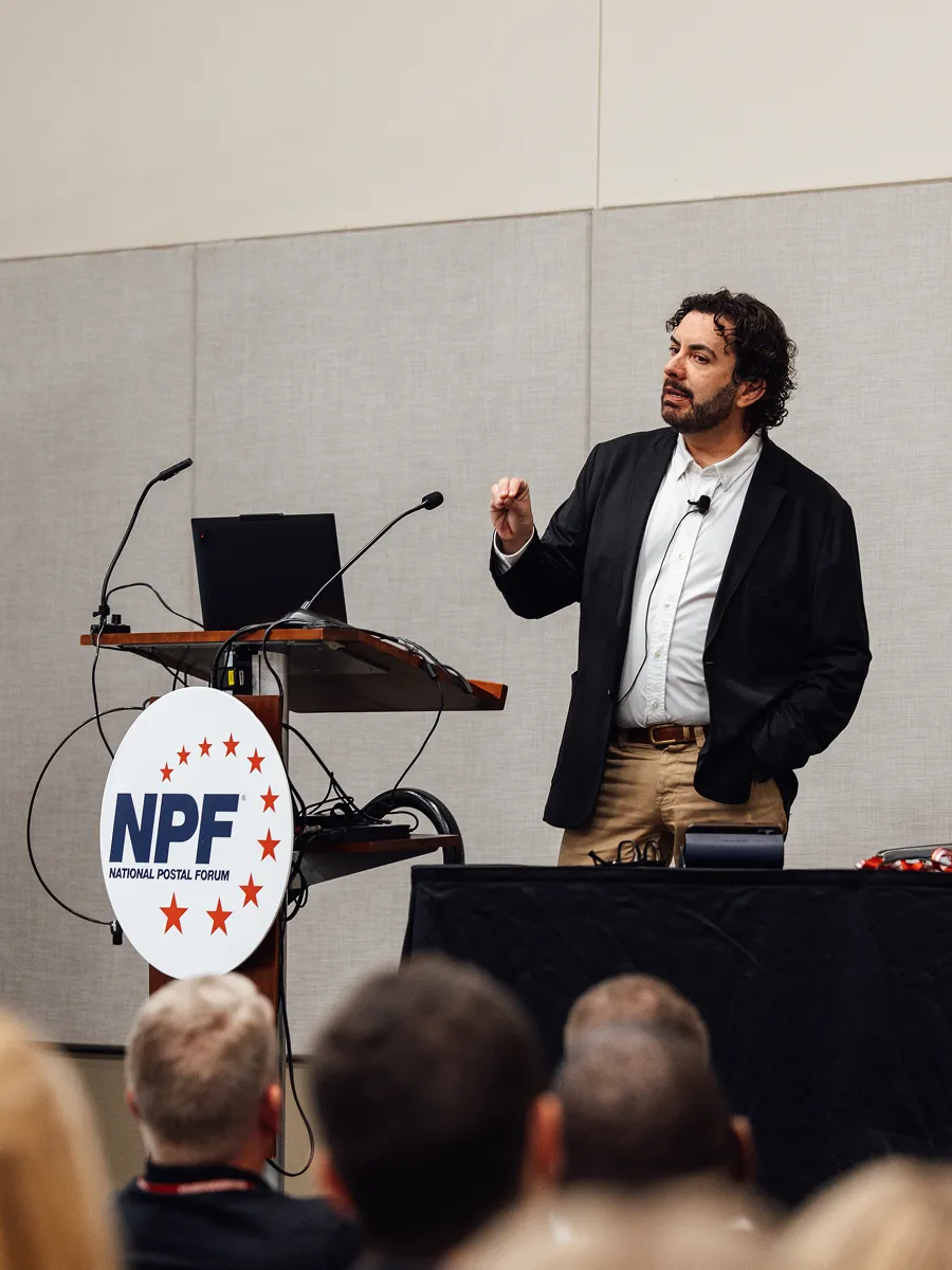 Man in blazer speaking at National Postal Forum with audience and podium in view.