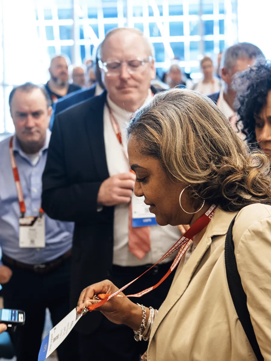 Woman reading an event badge on a red lanyard with people in business attire behind her.