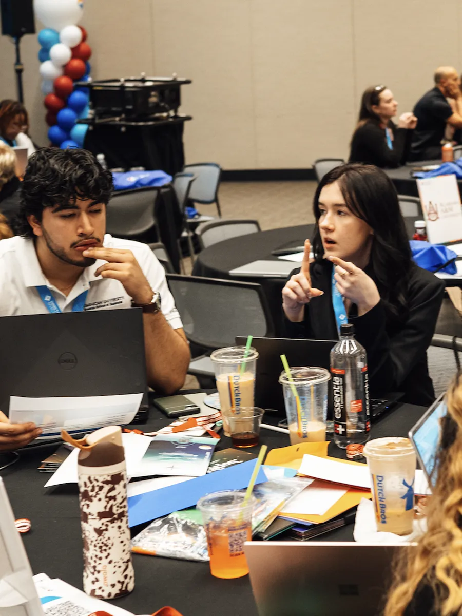 Two people seated at a table with laptops, papers, and drinks, engaged in discussion during a meeting.
