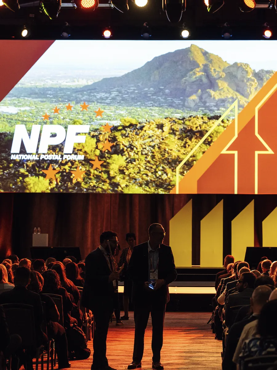 Audience seated in a dark conference room with a large screen showing NPF National Postal Forum logo and landscape.