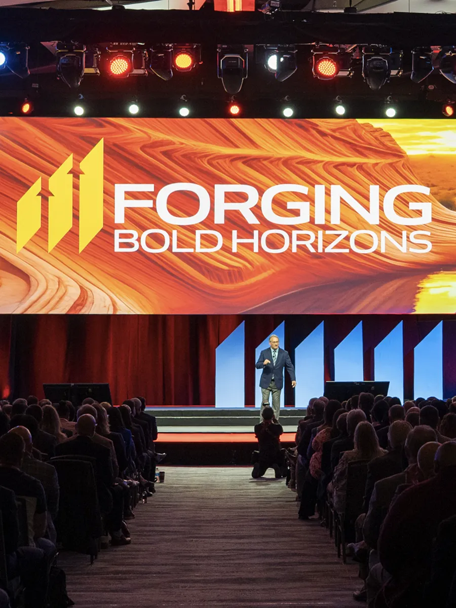 Speaker on stage at conference with large screen reading 'Forging Bold Horizons' and audience seated.