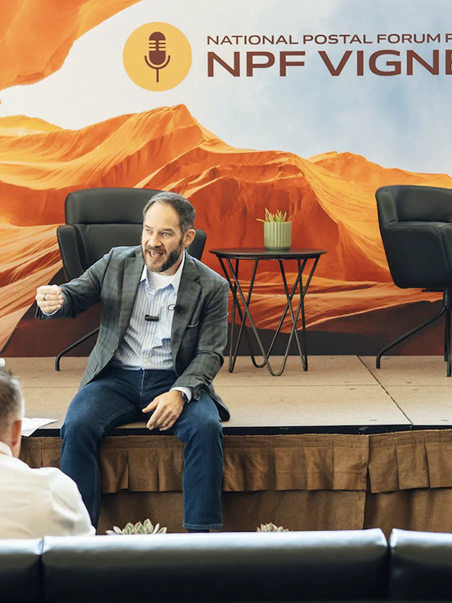 Man in a checkered blazer speaking animatedly on stage in front of a desert landscape backdrop.