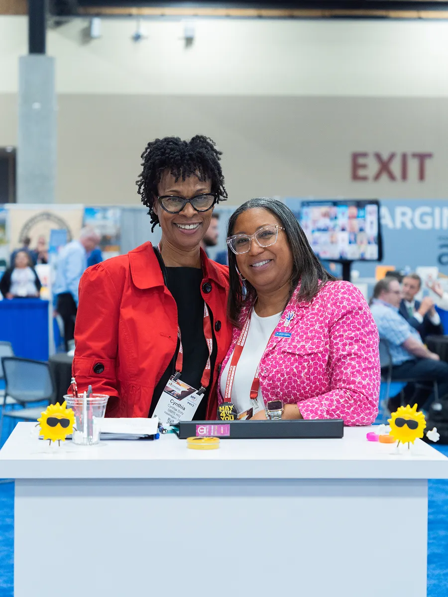 Two smiling women standing behind a booth table at a conference, one in red jacket, one in pink.