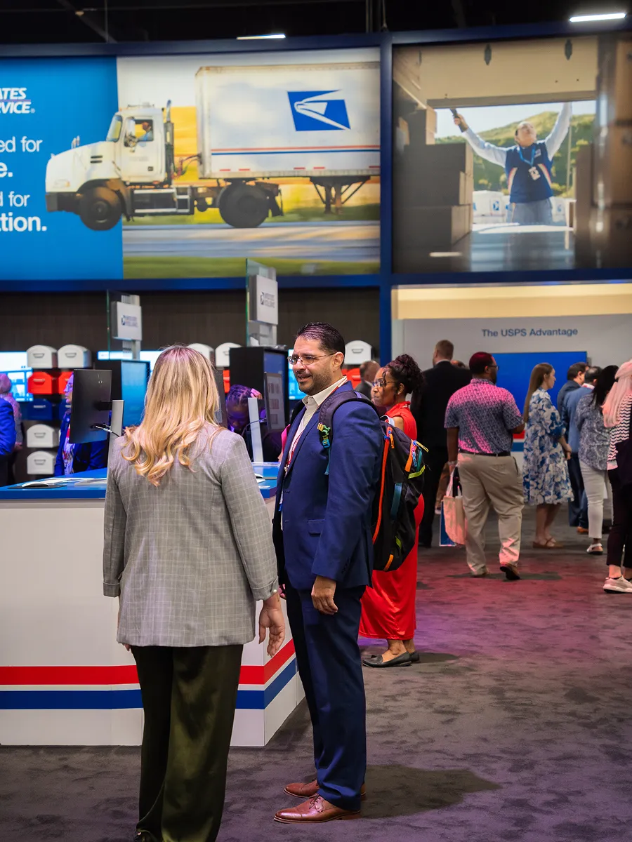 People talking and walking near USPS service counters inside a busy event or exhibition hall.