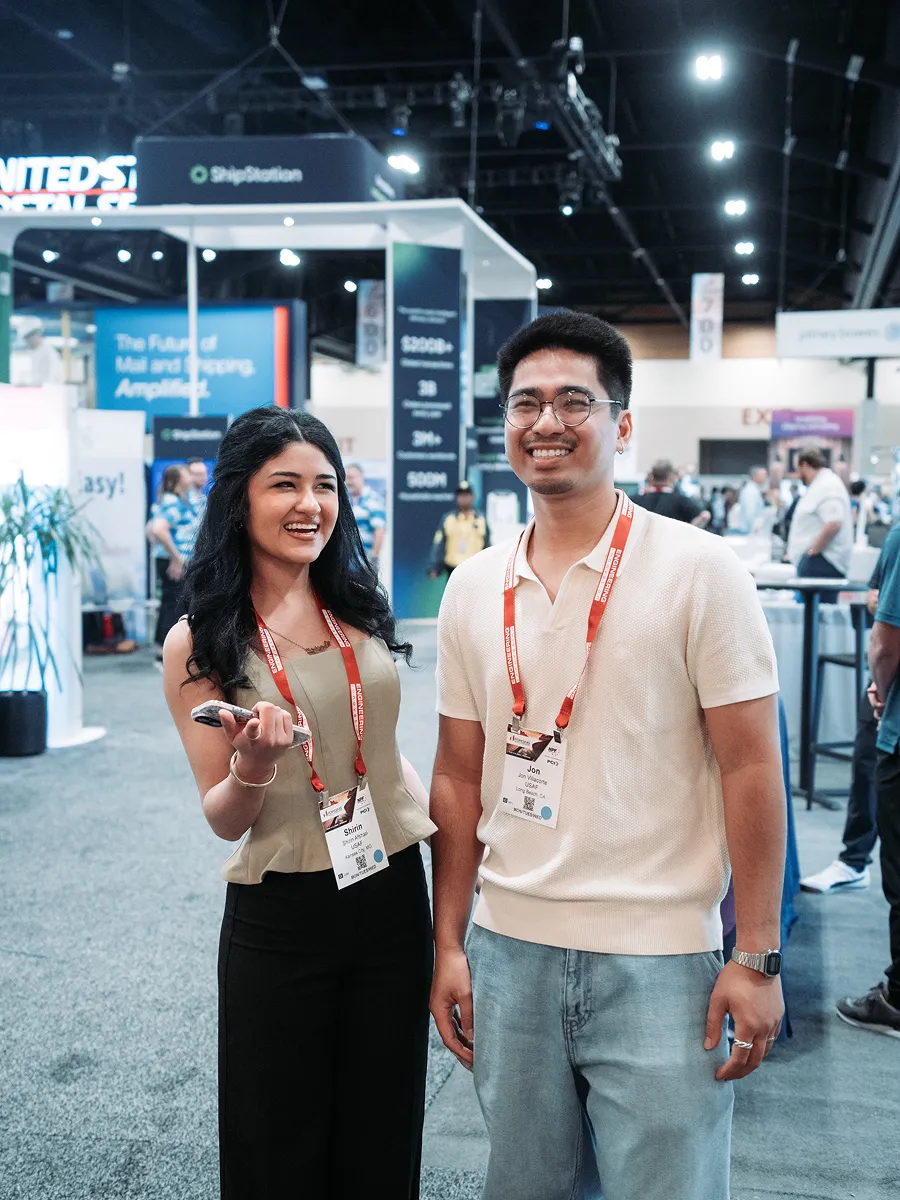 Two convention attendees smiling, wearing name badges and red lanyards in an exhibition hall.