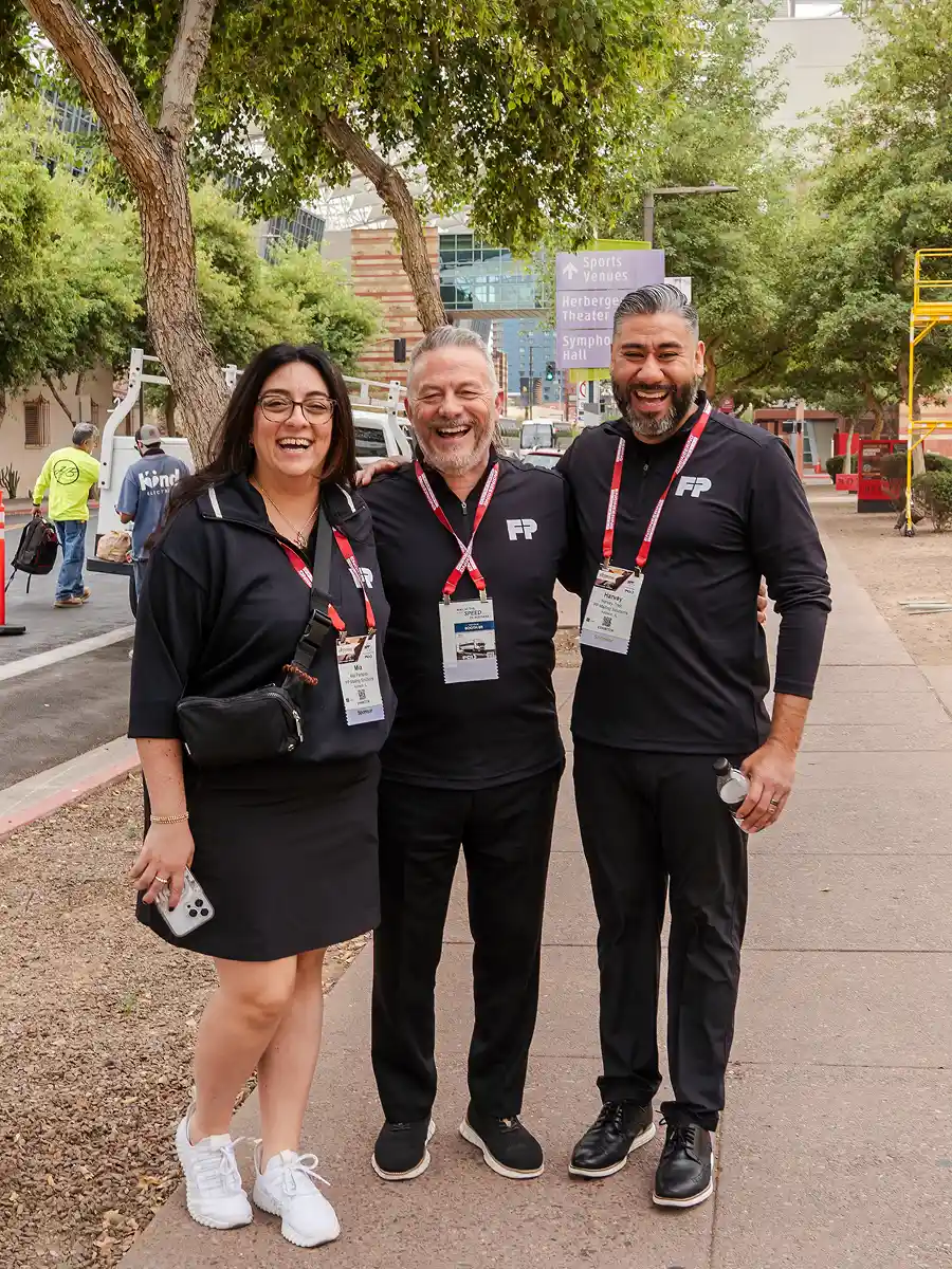 Three smiling people wearing black shirts with FP logo and event badges standing on a tree-lined sidewalk.