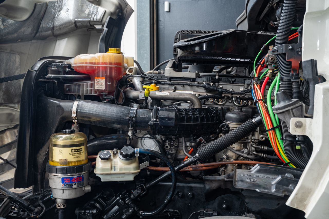 Open truck hood showing diesel engine components, coolant reservoir, fuel filter, hoses, and wiring during maintenance inside a professional repair shop.