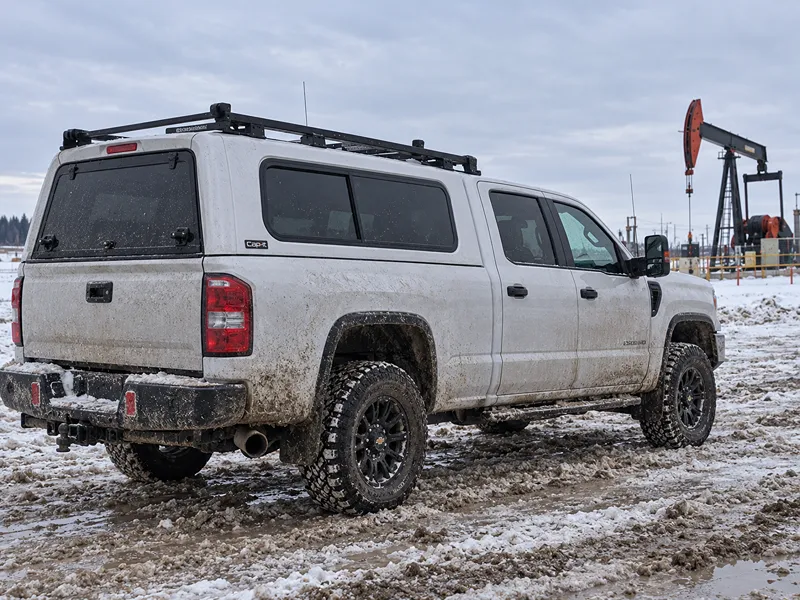 Silver pickup with a black V CANOPY truck cap, roof rack, and off-road tires parked in snow near an oil pumpjack.