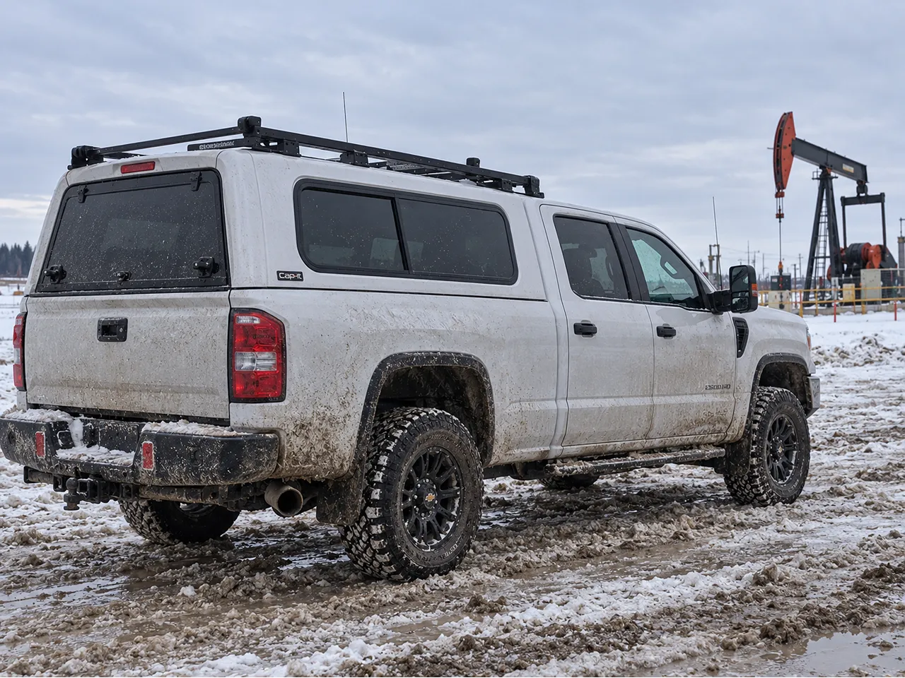 Silver pickup with a black V CANOPY truck cap, roof rack, and off-road tires parked in snow near an oil pumpjack.