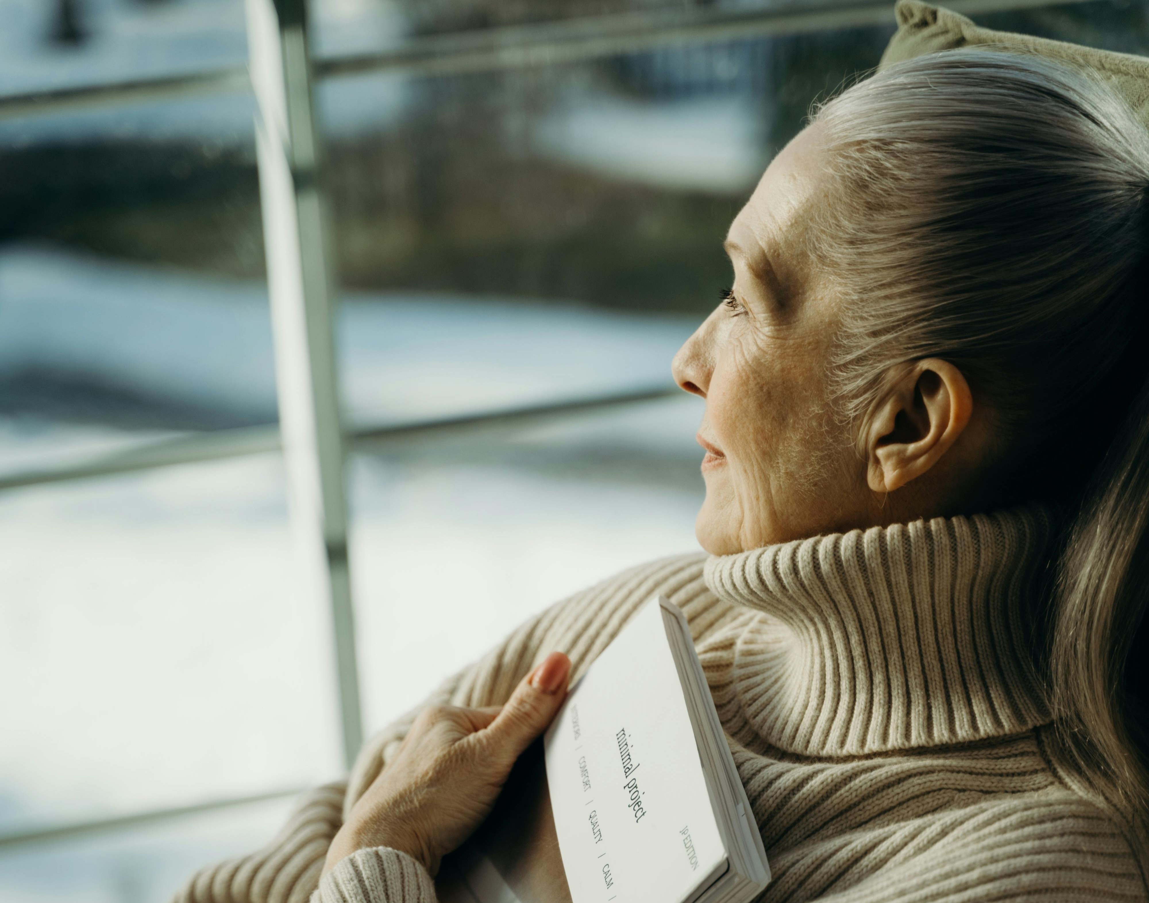 Older woman in a turtleneck gazing out a window with a thoughtful expression.