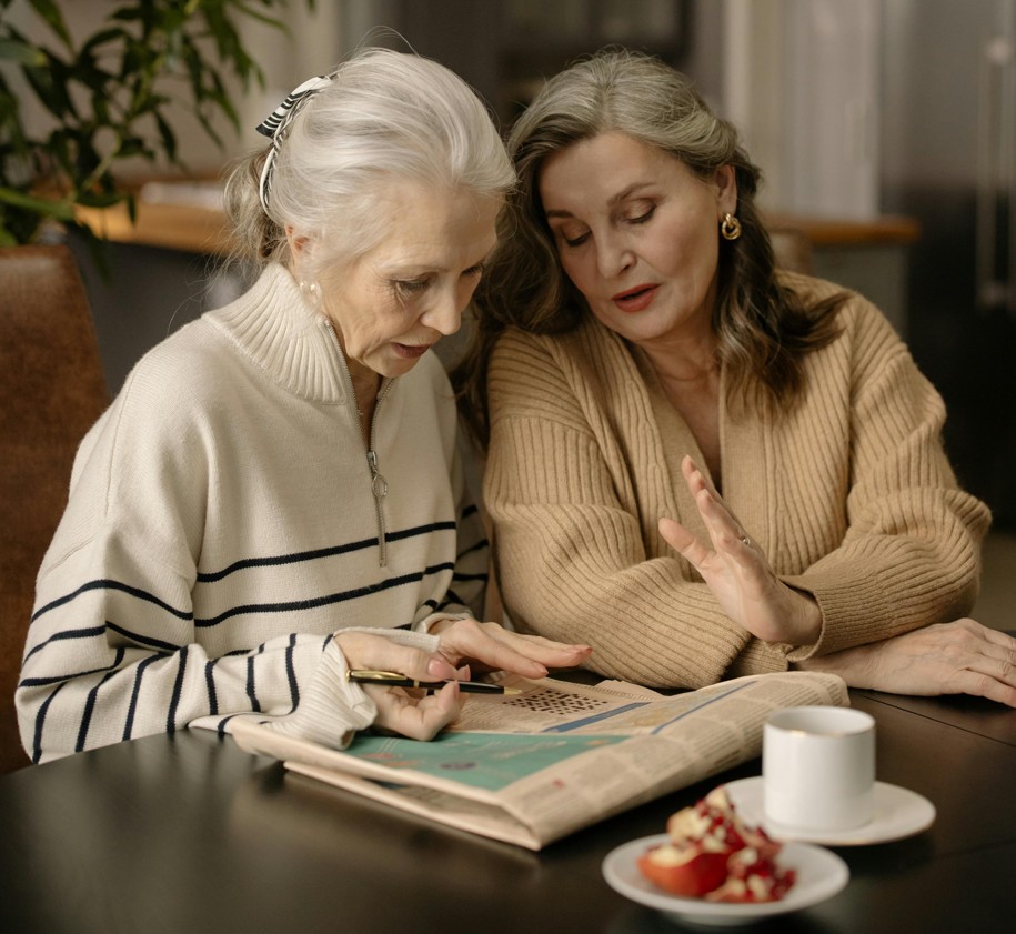 Two older women reading and discussing a book together at a table.