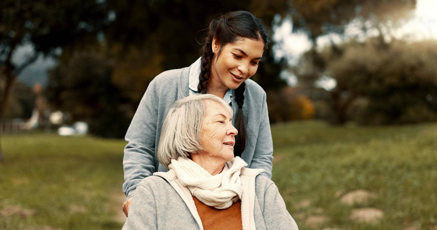 Caregiver standing behind a smiling older woman seated outdoors in a park.