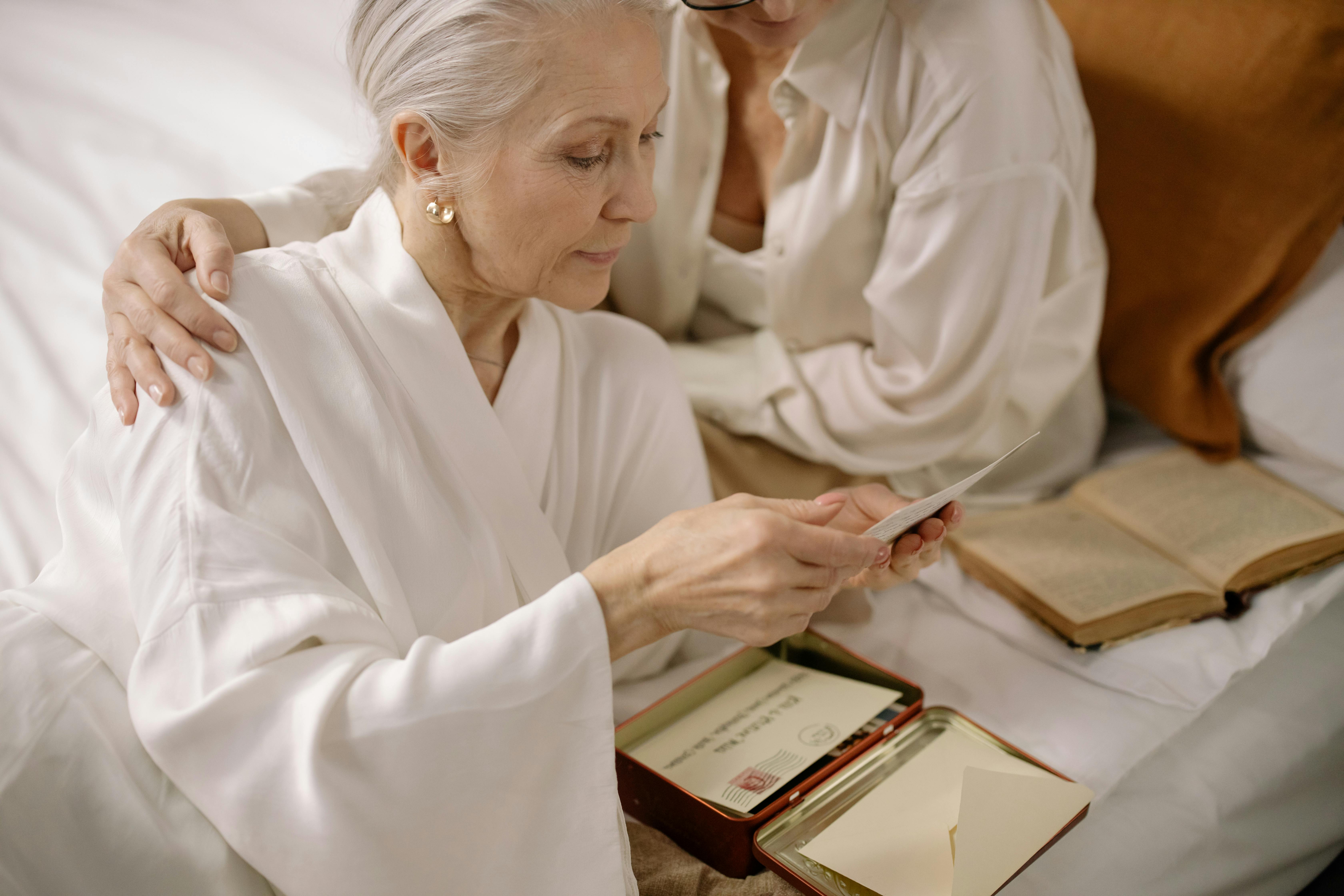 Older woman in a robe looking through old letters while a caregiver comforts her.