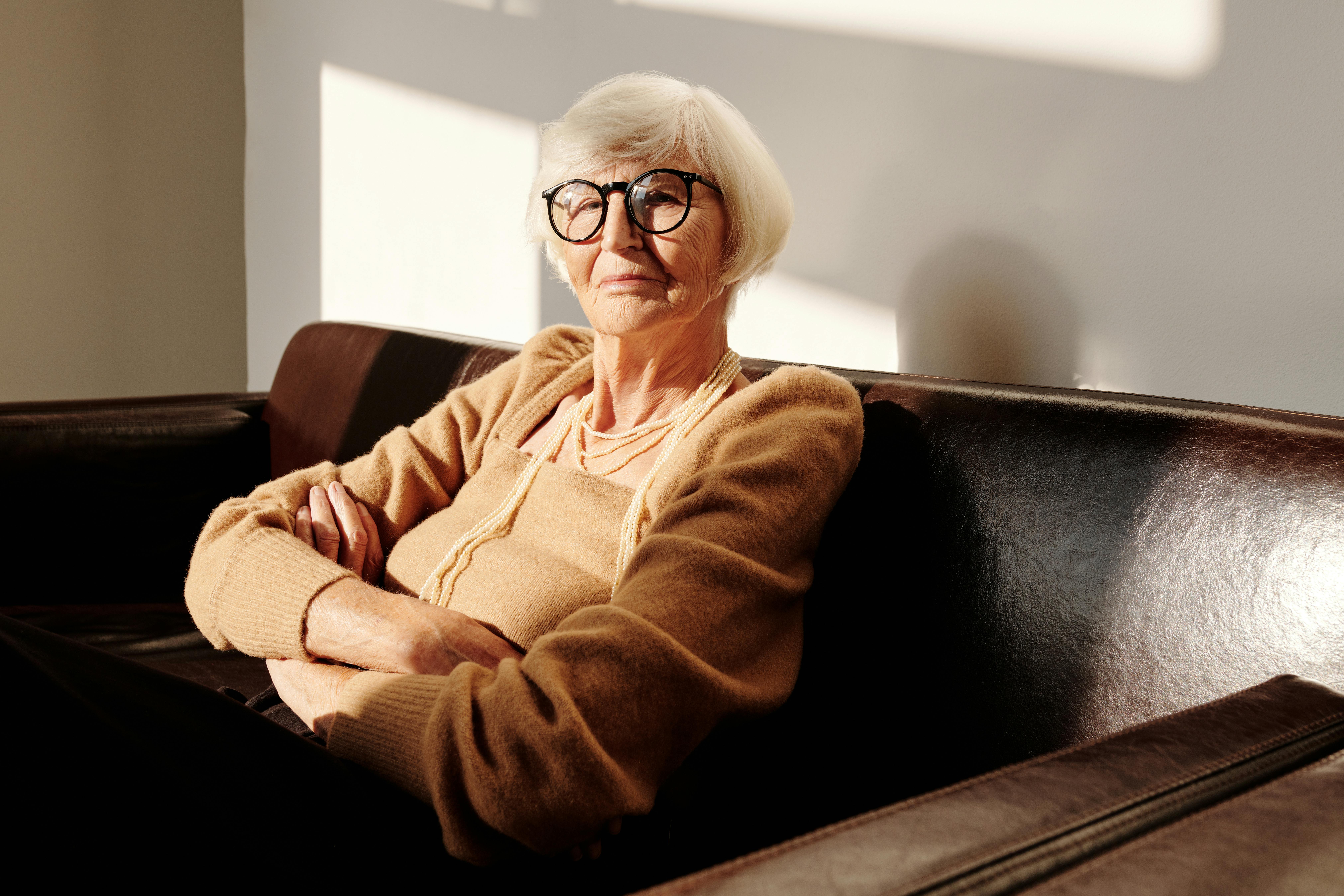 Older woman in glasses sitting on a sofa with her arms crossed and a serious expression.