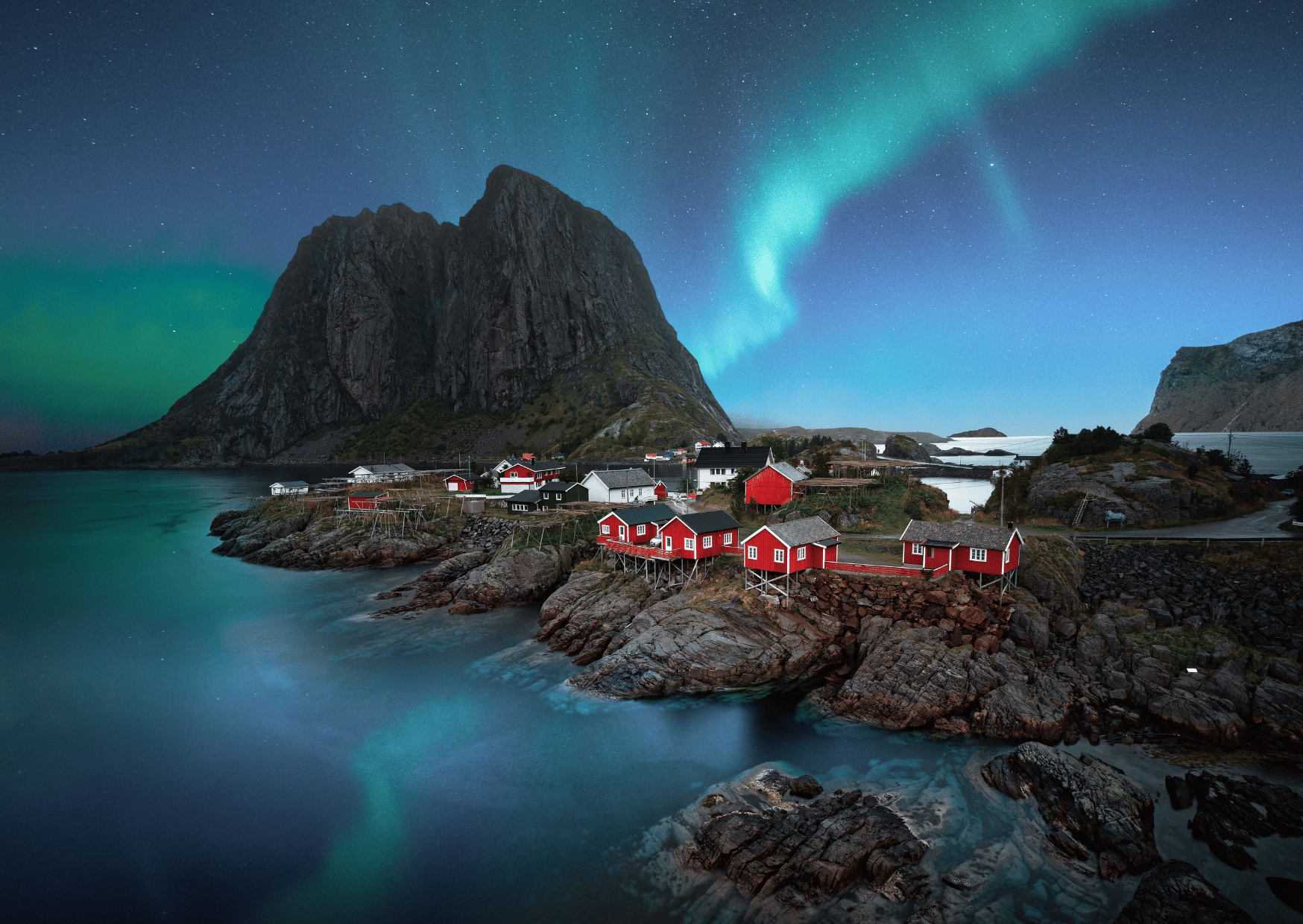 A photo from Lofoten showing northern lights over mountains and a small village with wooden houses