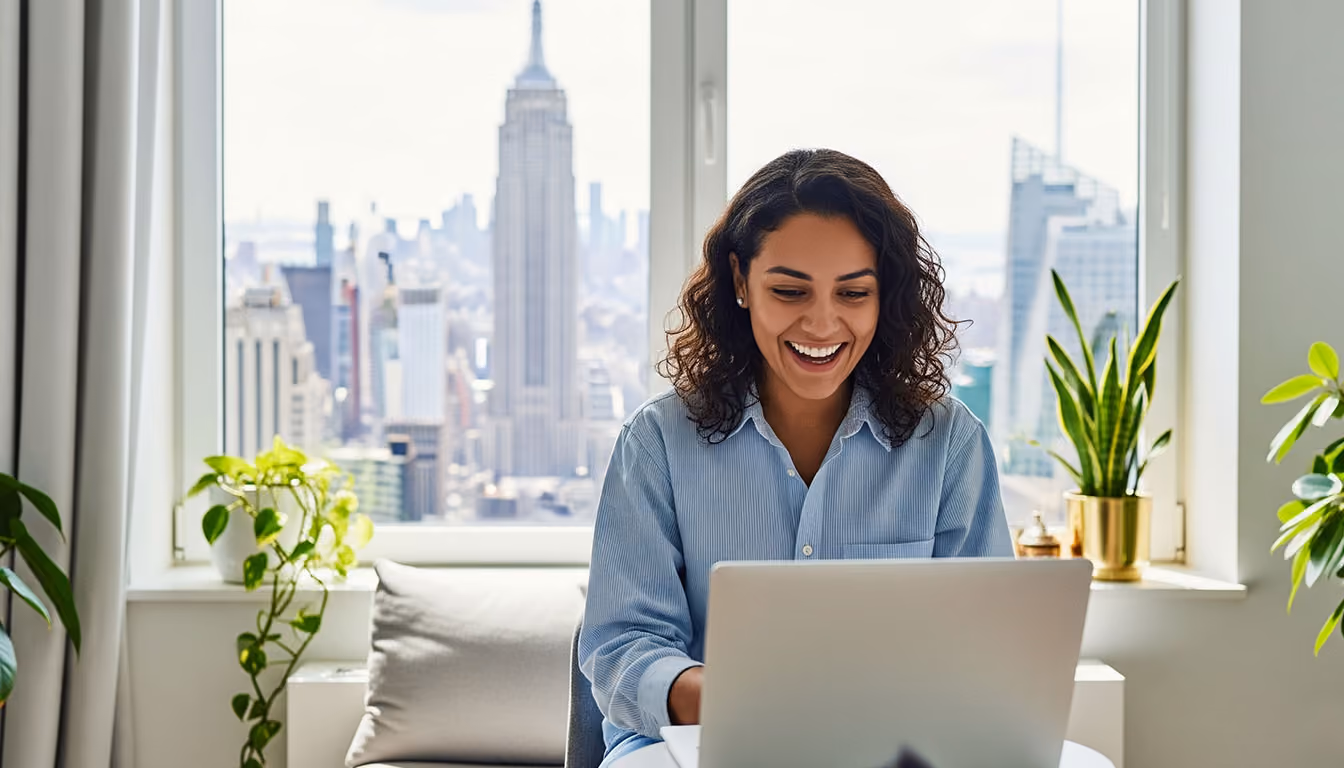 Smiling woman using laptop at desk with city skyline, including the Empire State Building, visible through window behind her.