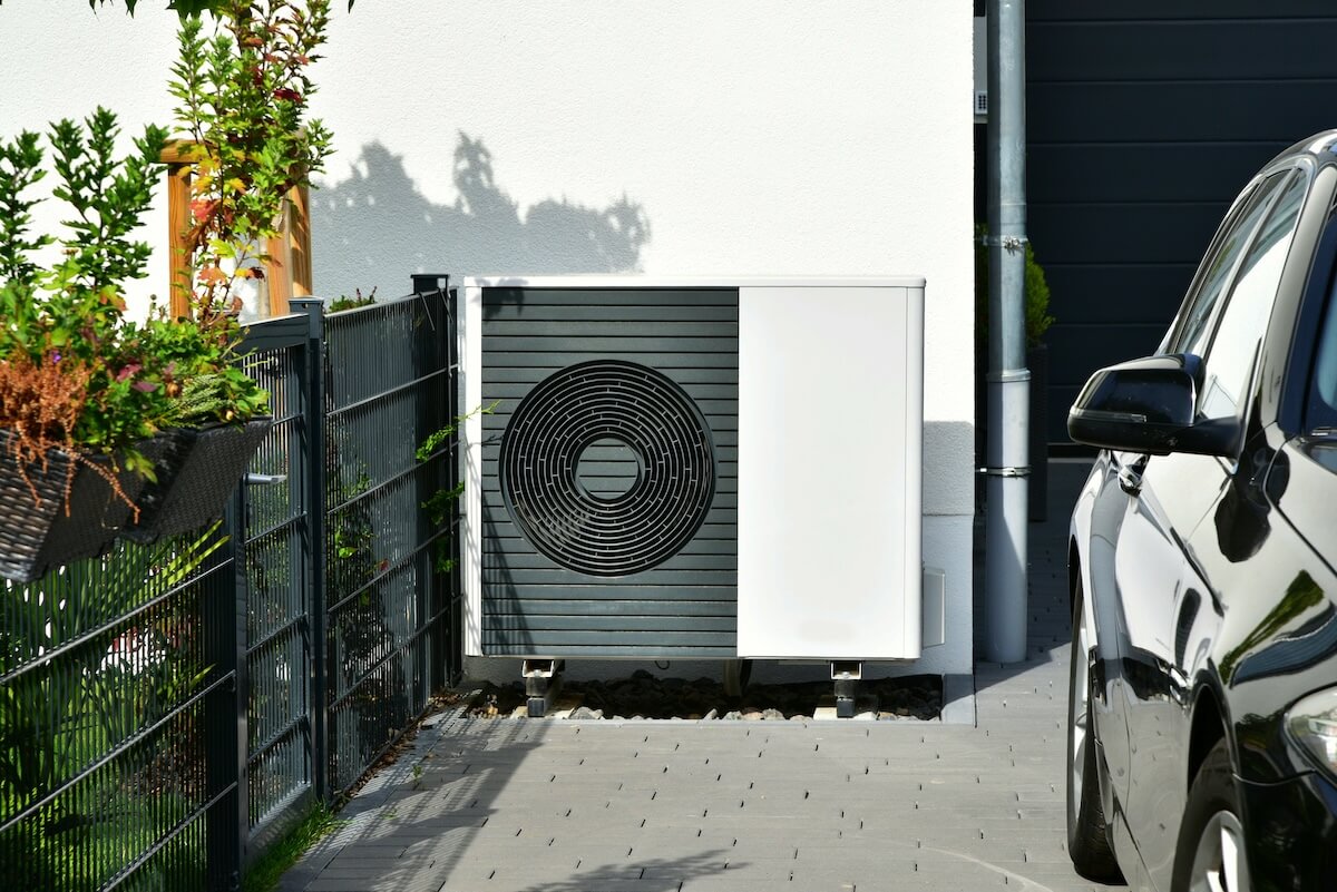 Wärmepumpe an der Hauswand in der Einfahrt eines Hauses zwischen Zaun zum Vorgarten und Auto