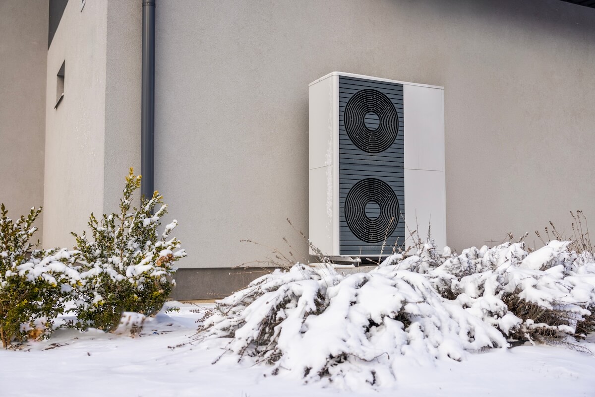 Wärmepumpe an Hauswand hinter schneebedeckten Büschen