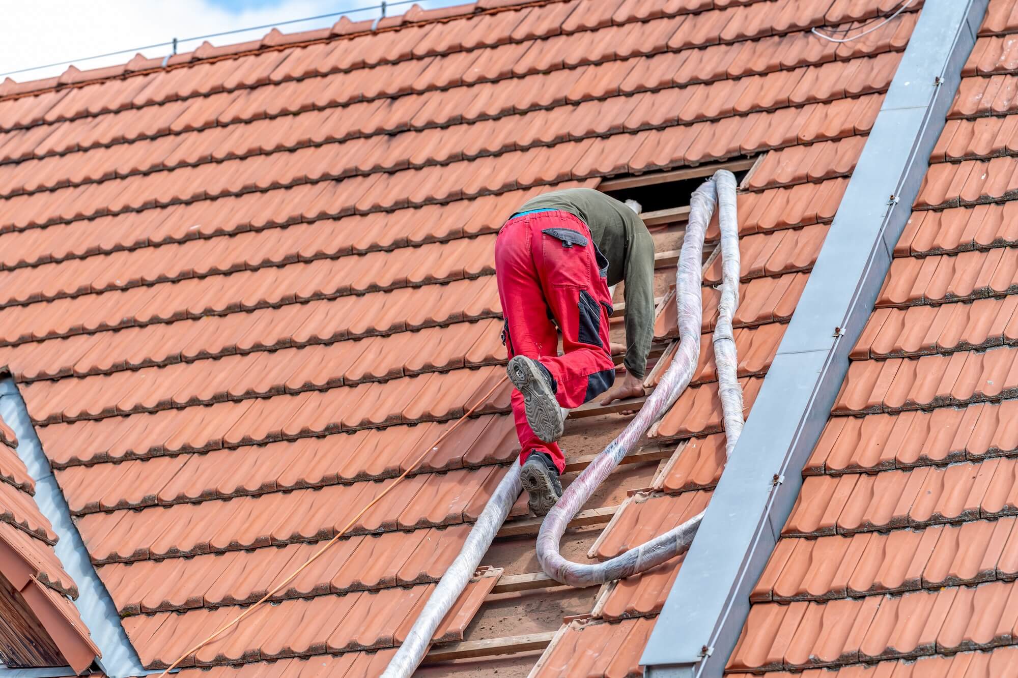 Ein Handwerker klettert auf einem Dach zu einem für die Einblasdämmung freigelegten Loch in den Ziegeln.