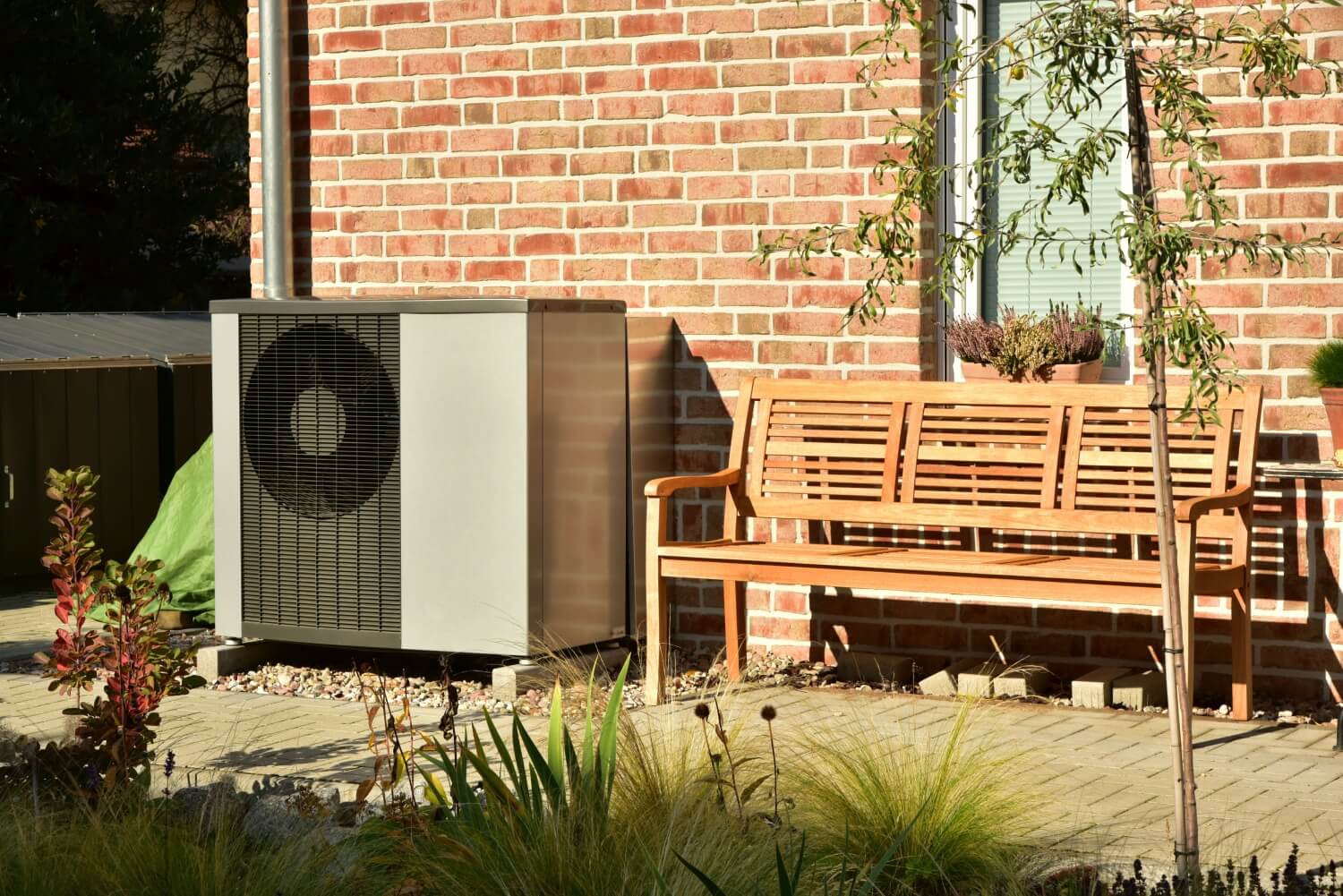 Graue Außeneinheit einer Wärmepumpe steht auf einem Fundament aus Beton vor einem roten Backsteinhaus