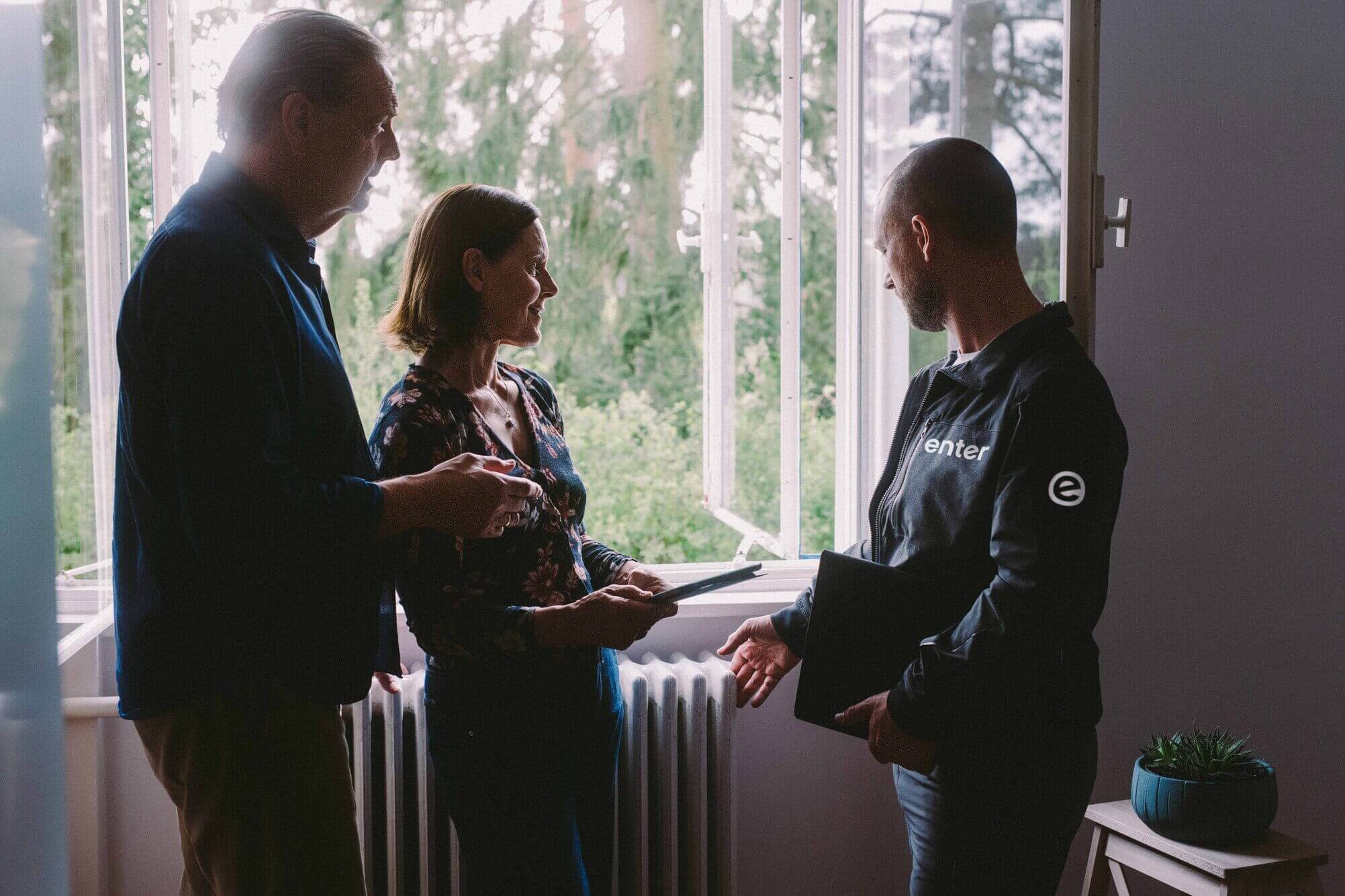 Ein Energieberater von Enter bespricht mit einem älteren Ehepaar die energetische Situation ihres Hauses am geöffneten Fenster.