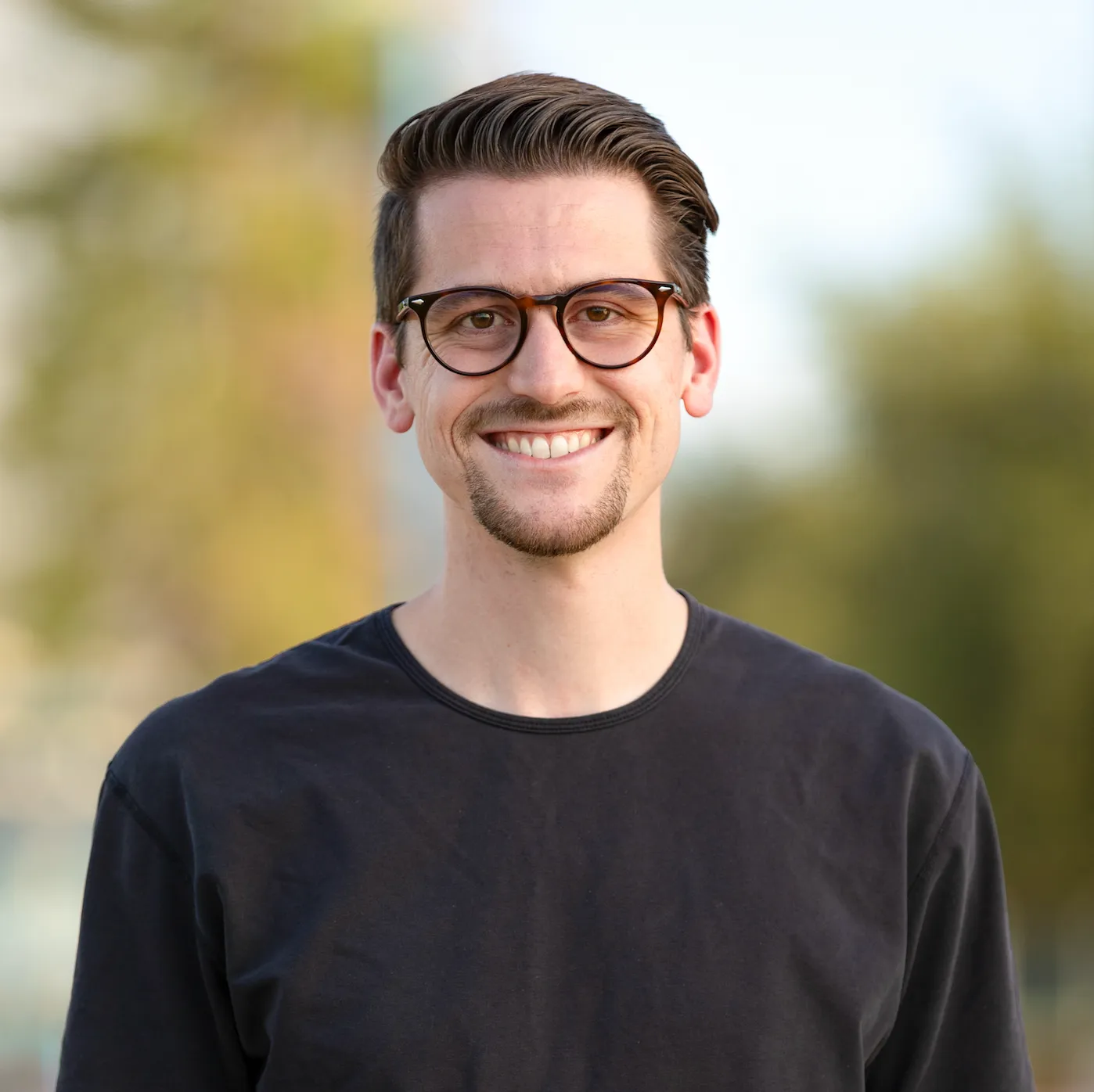 Smiling man with glasses and neatly styled hair wearing a black shirt outdoors.