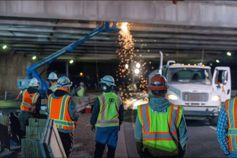 Construction crew in safety vests and hard hats watching sparks fly from overhead welding work on a bridge or highway overpass at night. A white truck is visible in the background.