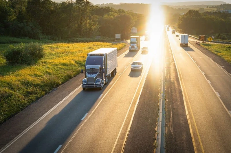 View of a sunny highway with a dark semi-truck driving in the foreground and other traffic, including cars and trucks, traveling in both directions.