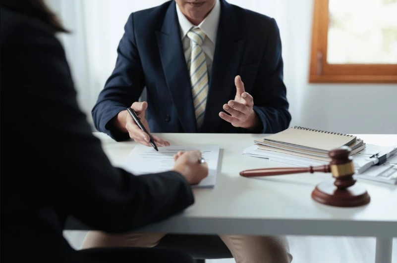 Close-up of a client signing a legal document on a white desk while a lawyer in a dark suit and tie gestures with a pen. A wooden gavel and stacked notebooks are visible on the desk.