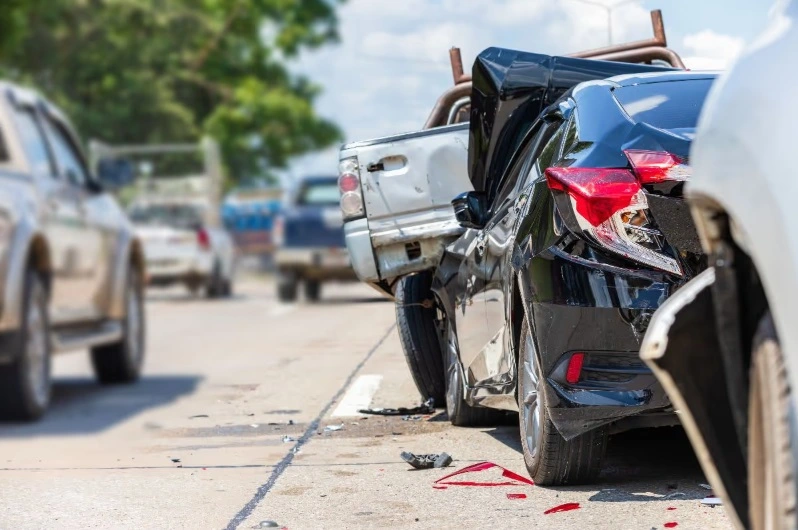Close-up view of a multi-car collision on a bright highway, showing rear-end damage to a black sedan, a silver pickup truck, and a white vehicle in the foreground, with red fluid on the road.