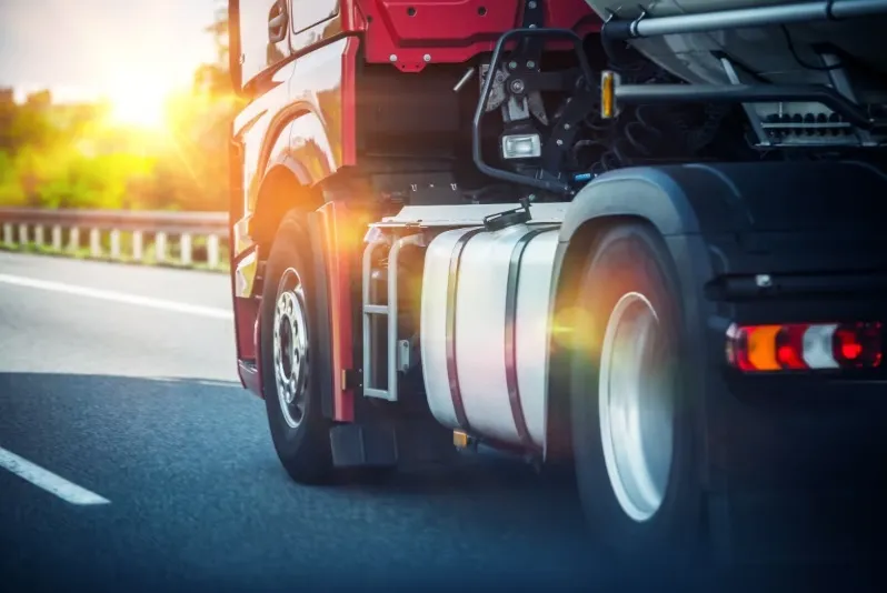 Rear view of a red semi-truck or transport truck driving fast on a sunny asphalt highway with a bright sun flare over the cab.
