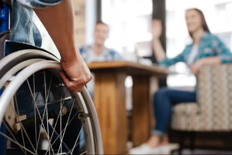 Close-up on a person's hand gripping the wheel of a wheelchair, with two friends seated at a table and waving in the background, suggesting an accessible social setting like a cafe.