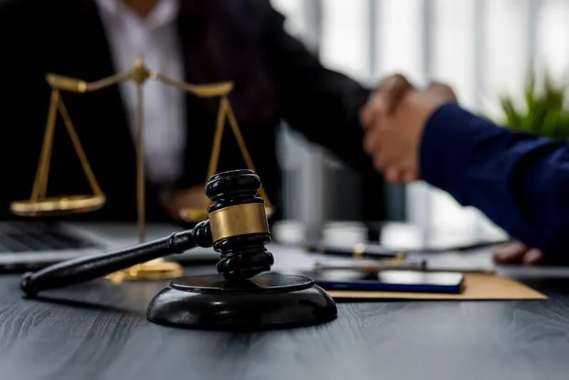 Close-up of a wooden judge's gavel and the brass scales of justice on a desk, with two people shaking hands in the blurred background, signifying a legal agreement or settlement.