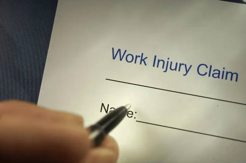 Close-up of a hand with a pen pointing to the 'Name' line on a document titled "Work Injury Claim," indicating the start of filing a workers' compensation application.