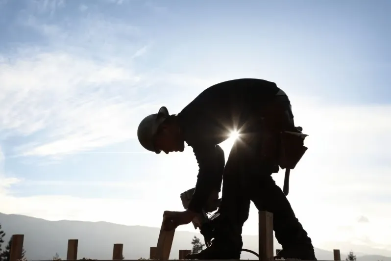 Silhouette of a construction worker or carpenter wearing a hard hat and tool belt, using a power drill to assemble a wood frame against a bright sky at sunrise or sunset.