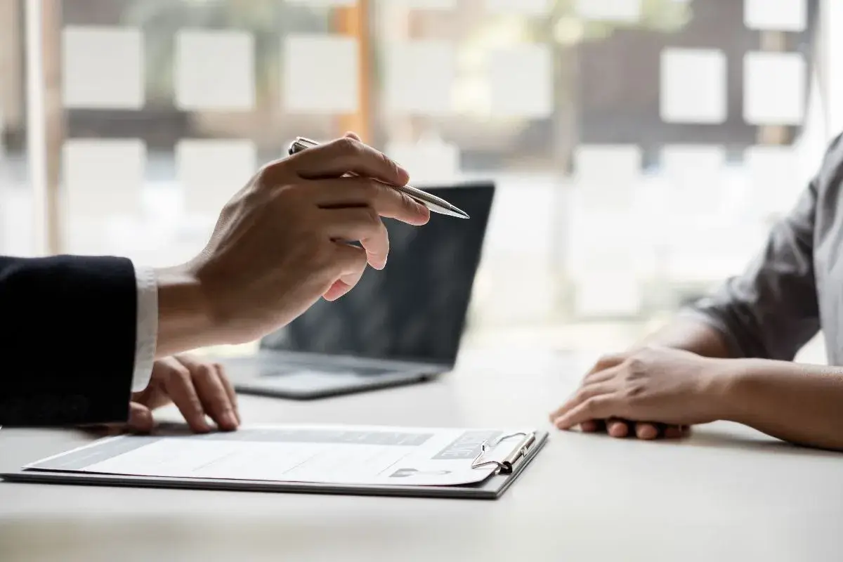 Close-up of a person's hands holding a pen and pointing to a document on a desk, possibly during a job interview, business consultation, or review of a resume. A blurred person and laptop are in the background.