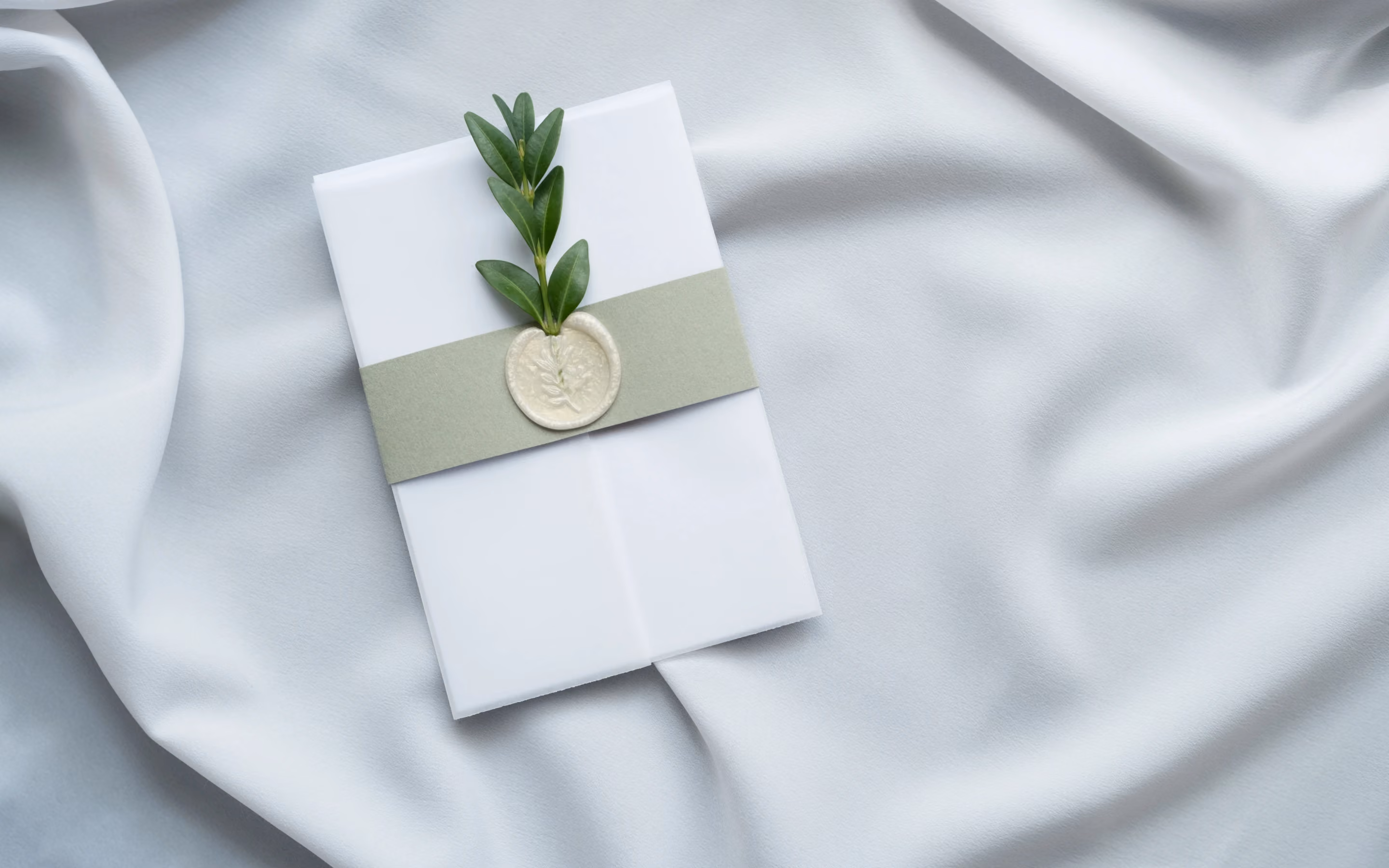 Elegant wedding invitation with botanical wax seal and olive branch on silk fabric background.